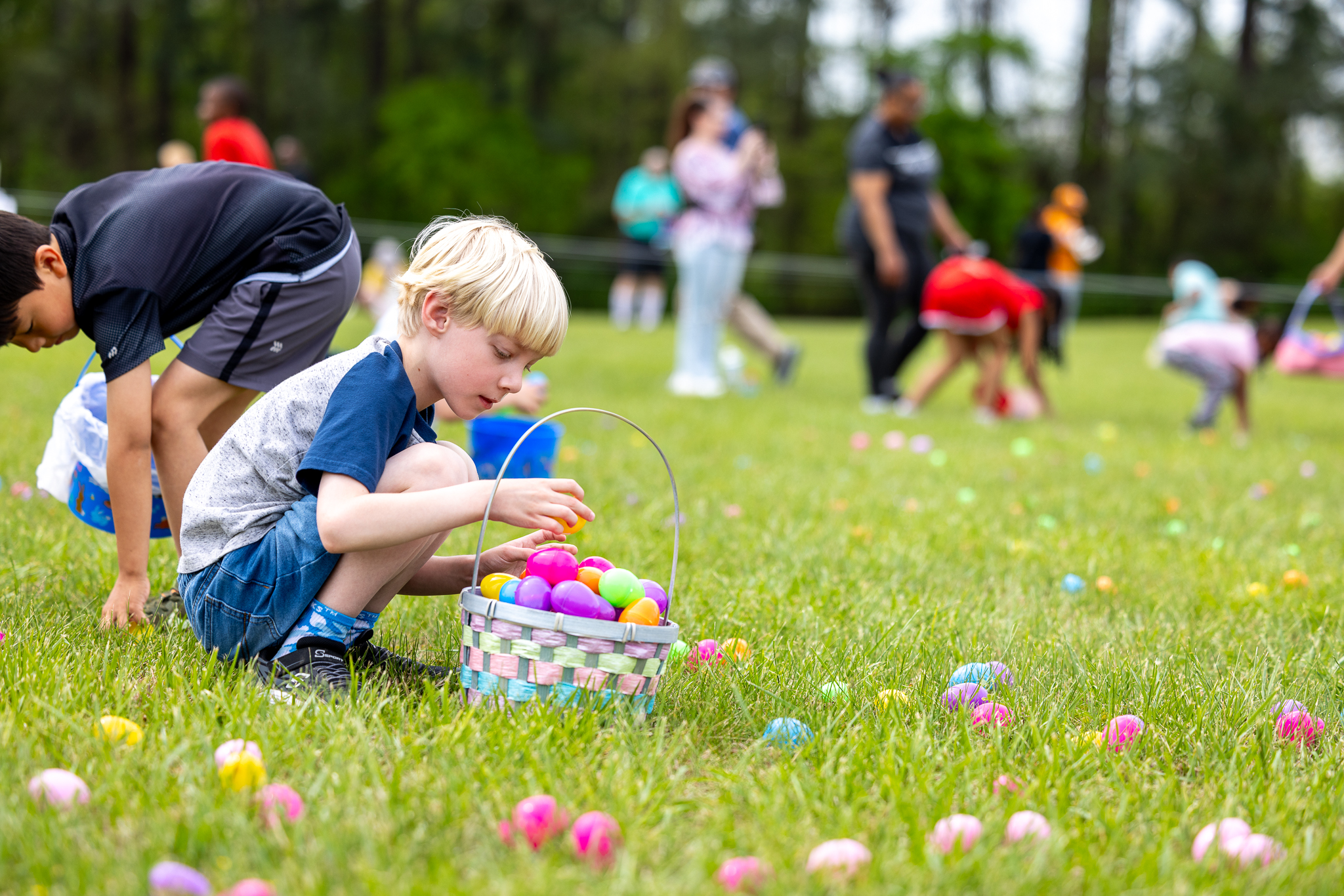 Photo of a young child crouched down picking up easter eggs during an easter egg hunt with an overflowing basket of eggs.